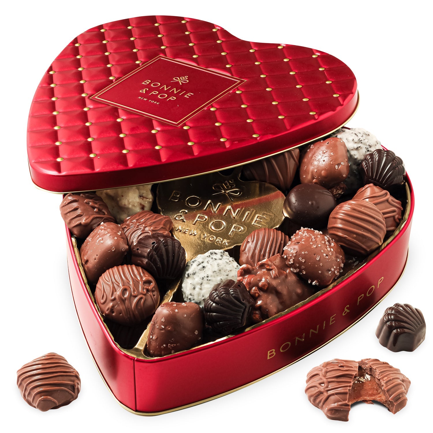 A red heart-shaped tin filled with an assortment of chocolates and chocolate-covered snacks, including truffles and pretzels, with a decorative pattern on the lid.