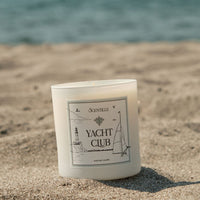 Candle labeled 'Yacht Club' on a sandy beach with ocean in the background