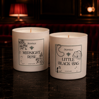 Two Scentille scented candles in white glass jars, labeled 'Midnight Rose' and 'Little Black Bag', displayed on a dark marble surface with a warm, dimly lit background.