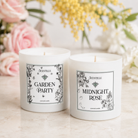 Two white Scentille scented candles, labeled 'GARDEN PARTY' and 'MIDNIGHT ROSE', displayed on a light marble surface with soft pink and yellow flowers blurred in the background.
