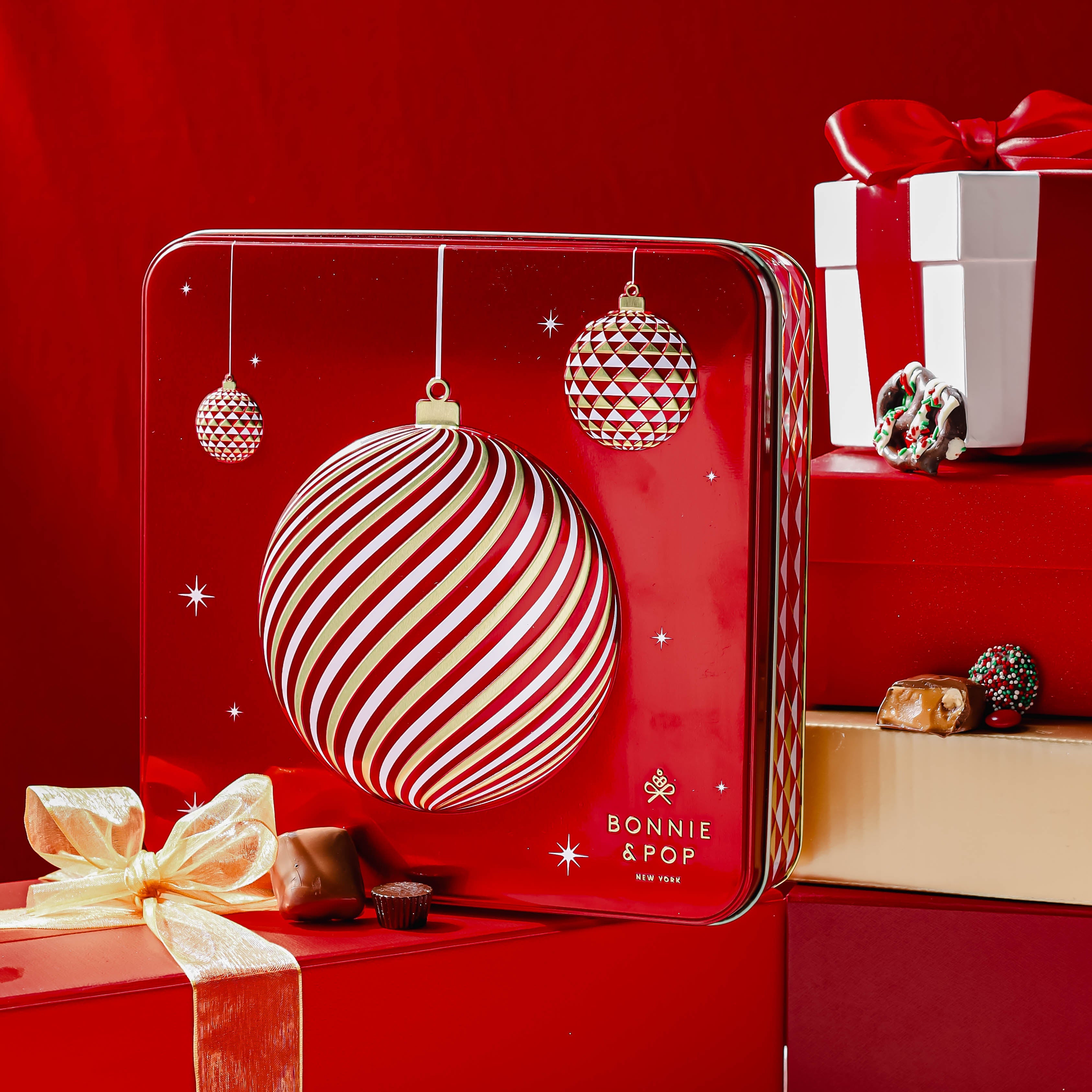 A red tin with Christmas-themed decorations containing an assortment of chocolate treats including milk chocolate covered Oreos, hazelnut truffles, and other holiday snack mixes.