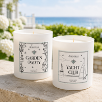 Two Scentille scented candles in white glass jars are displayed outdoors on a stone ledge with a blurred garden and ocean background. The left jar is labeled 'GARDEN PARTY' with botanical illustrations, and the right jar is labeled 'YACHT CLUB' with nautical line drawings.